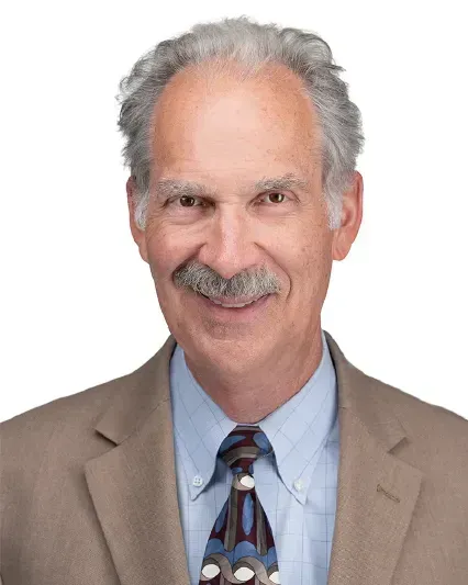 Portrait of a man in a tan suit, blue shirt, and patterned tie, smiling against a white background