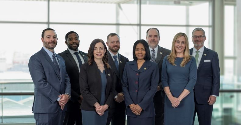 Seven professionals in business attire standing in a bright office lobby, posing for a group photo.