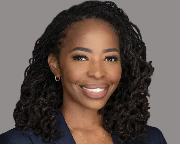 Portrait of a smiling woman with curly hair in a dark blazer against a gray background