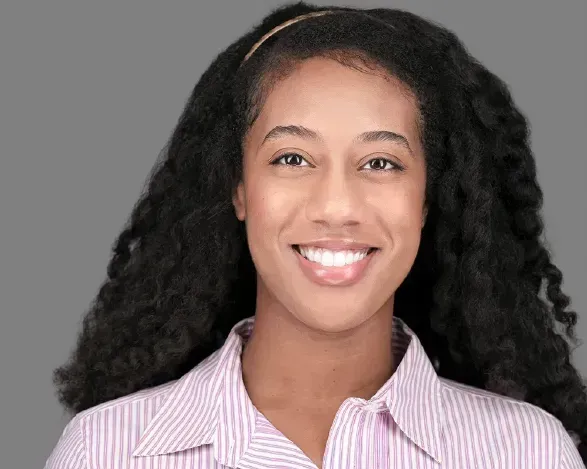 Smiling person with long curly hair wearing a light striped shirt against a gray background