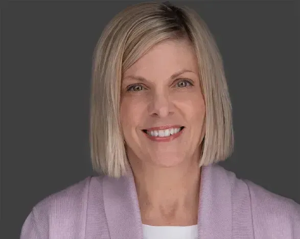 Smiling woman with a blonde bob wearing a light purple cardigan against a dark gray background