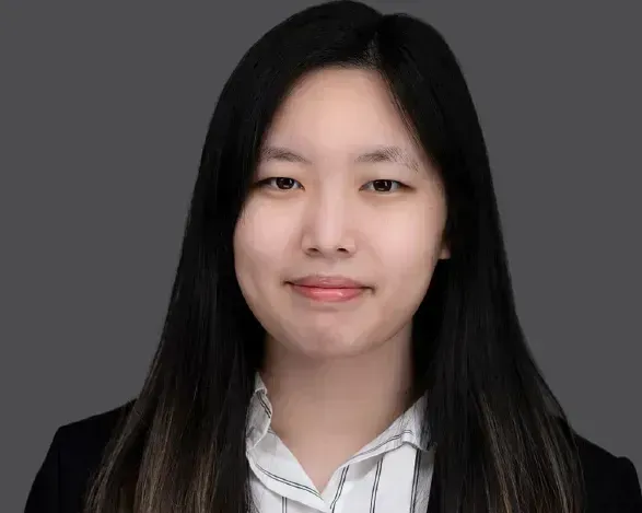 Headshot of a person with long black hair, wearing a white shirt and black blazer against a gray background