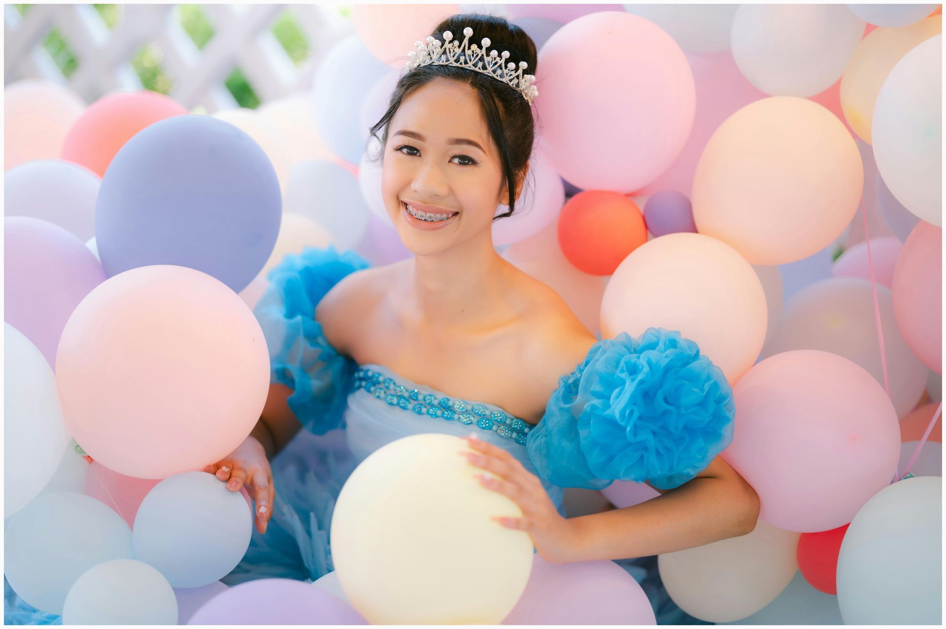 A woman in a blue dress and tiara is surrounded by balloons.