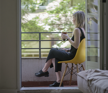 Uma mulher está sentada em uma cadeira amarela em uma varanda segurando uma xícara de café.