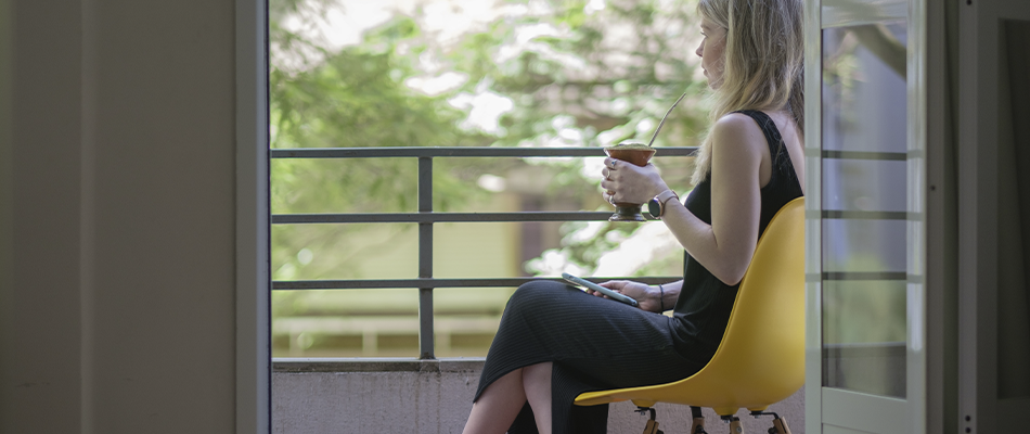 Uma mulher está sentada em uma cadeira amarela em uma varanda tomando café.