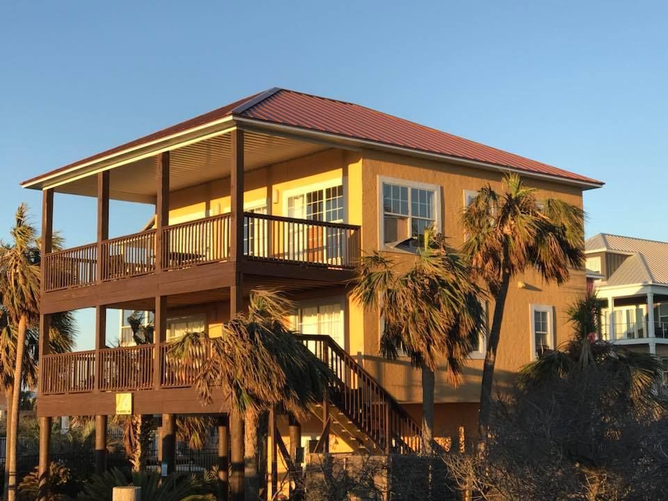 A two-story yellow stilt house with a reddish-brown metal roof and extensive wooden decks