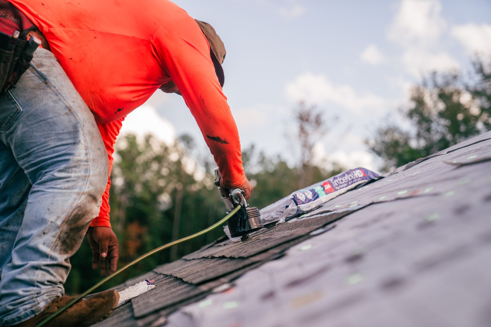 A roofer, wearing an orange safety shirt and a leather tool belt, is bent over and using a pneumatic