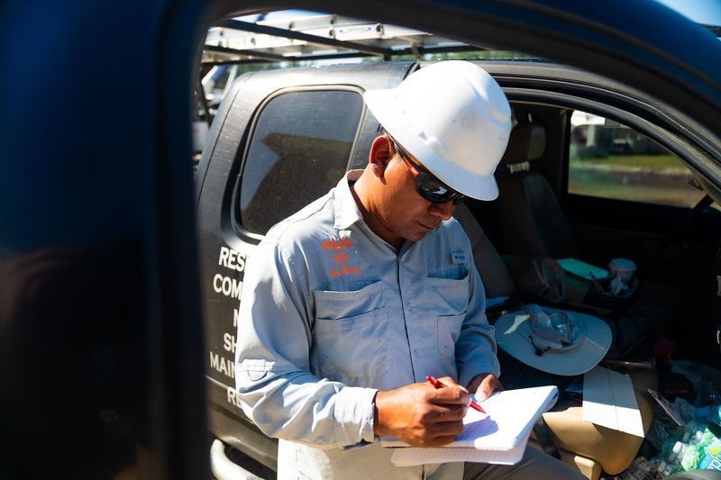 A roofing inspector wearing a white hard hat and safety glasses writes notes on a clipboard beside