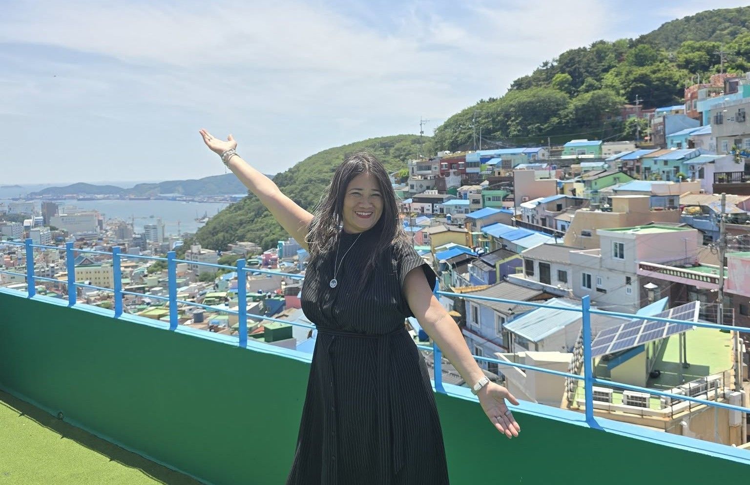 A woman in a black dress is standing on a balcony with her arms outstretched.