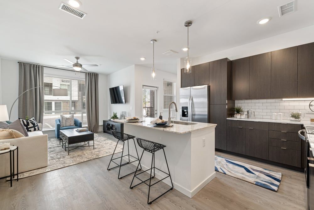 Modern open concept living room and kitchen area in an apartment with light wood flooring. The living room features a gray sofa, a blue armchair, a dark wood coffee table, and a television mounted on the wall. Large windows with gray curtains provide natural light. The kitchen is equipped with dark brown cabinets, white subway tile backsplash, stainless steel appliances, and a center island with a sink and bar stools. The space is accented with pendant lighting and decorative items, creating a cozy and stylish interior at Marq Perimeter in Atlanta, GA.