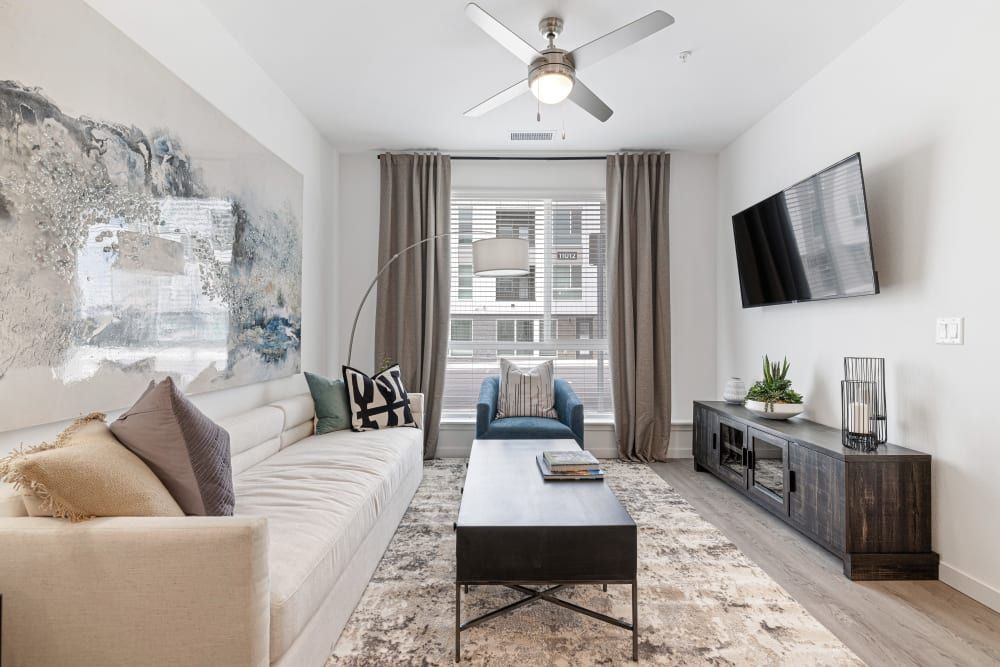 Modern living room with abundant natural light featuring a large sectional beige sofa adorned with decorative pillows, a blue armchair, a flat-screen TV mounted on the wall, and a dark wood entertainment console. The room is decorated with a large abstract painting, a cream area rug, a simplistic black coffee table, and floor-to-ceiling curtains on the windows. The space is completed with a ceiling fan, various decorative items, and potted plants creating a cozy and stylish interior at Marq Perimeter in Atlanta, GA.