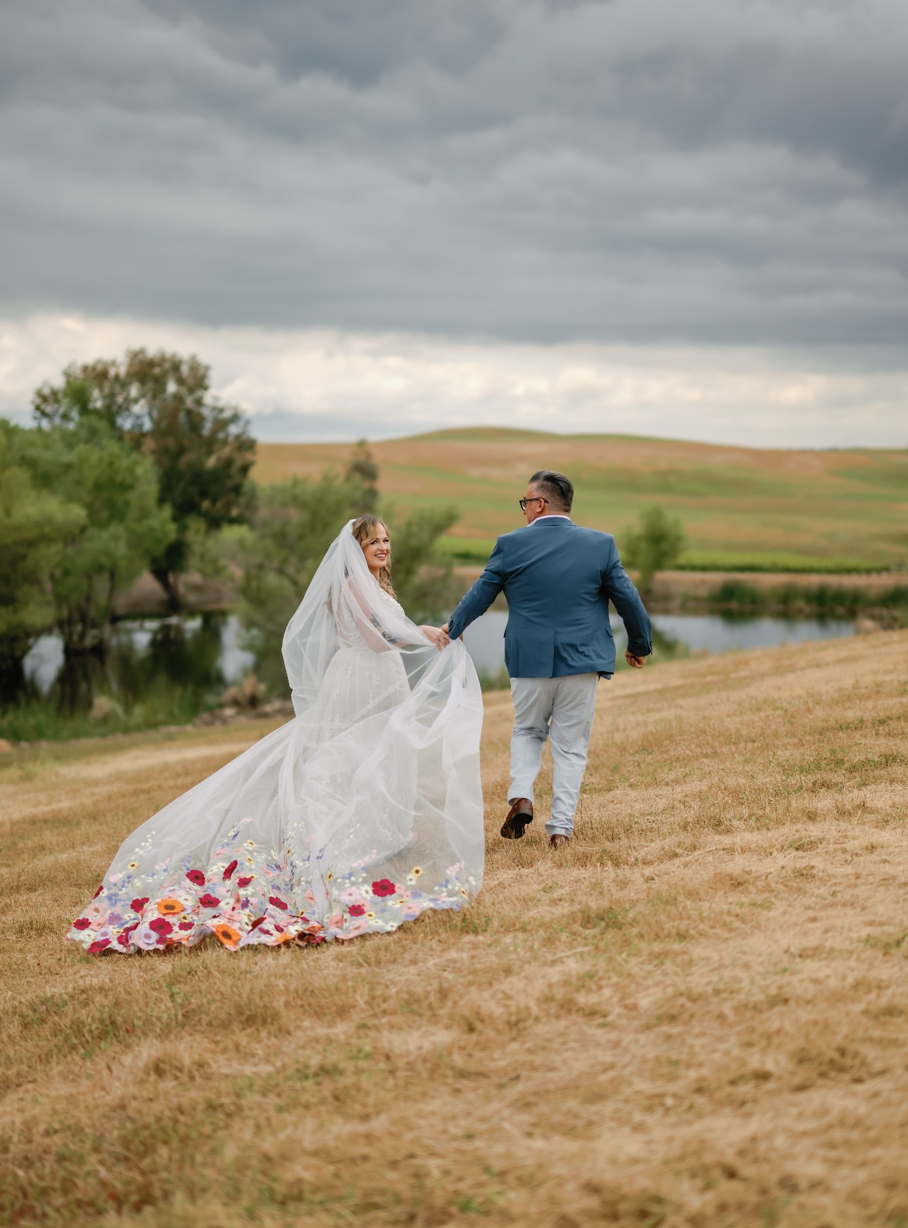 Bride and groom walk hand-in-hand across a field. The bride's gown has a long train with floral details, while the groom wears a blue jacket.