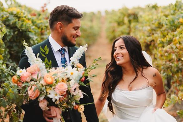 A bride and groom are walking through a vineyard holding hands and smiling.