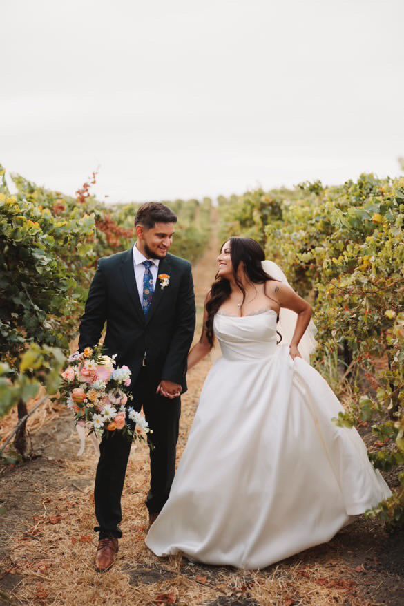 A bride and groom are walking through a vineyard holding hands.