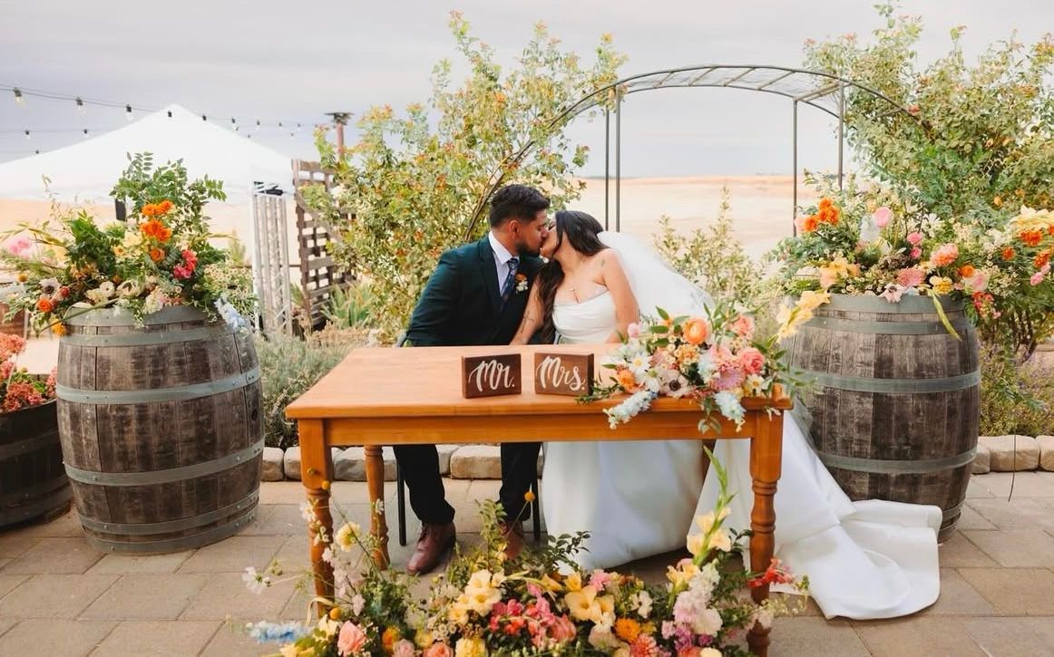 A bride and groom are kissing at a table at their wedding.
