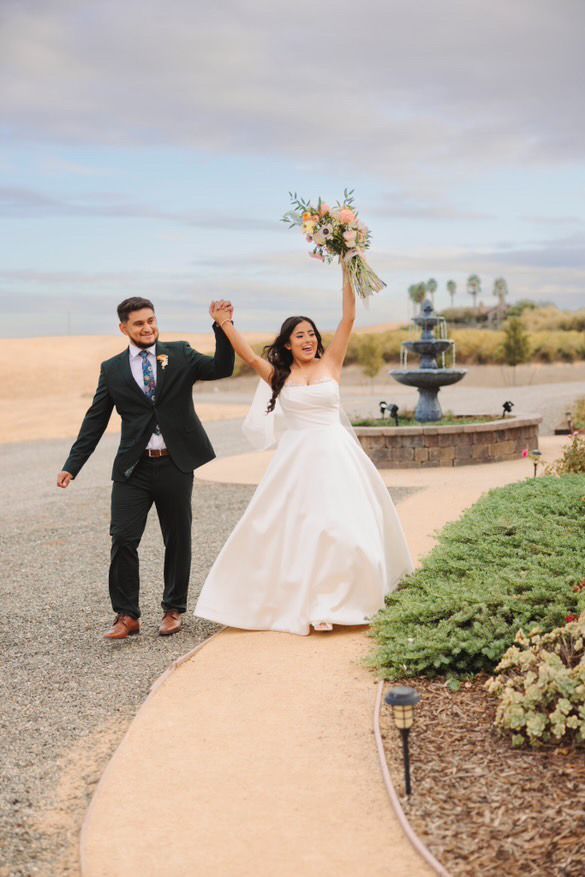 A bride and groom are walking down a path holding hands with a garden and fountain behind them.