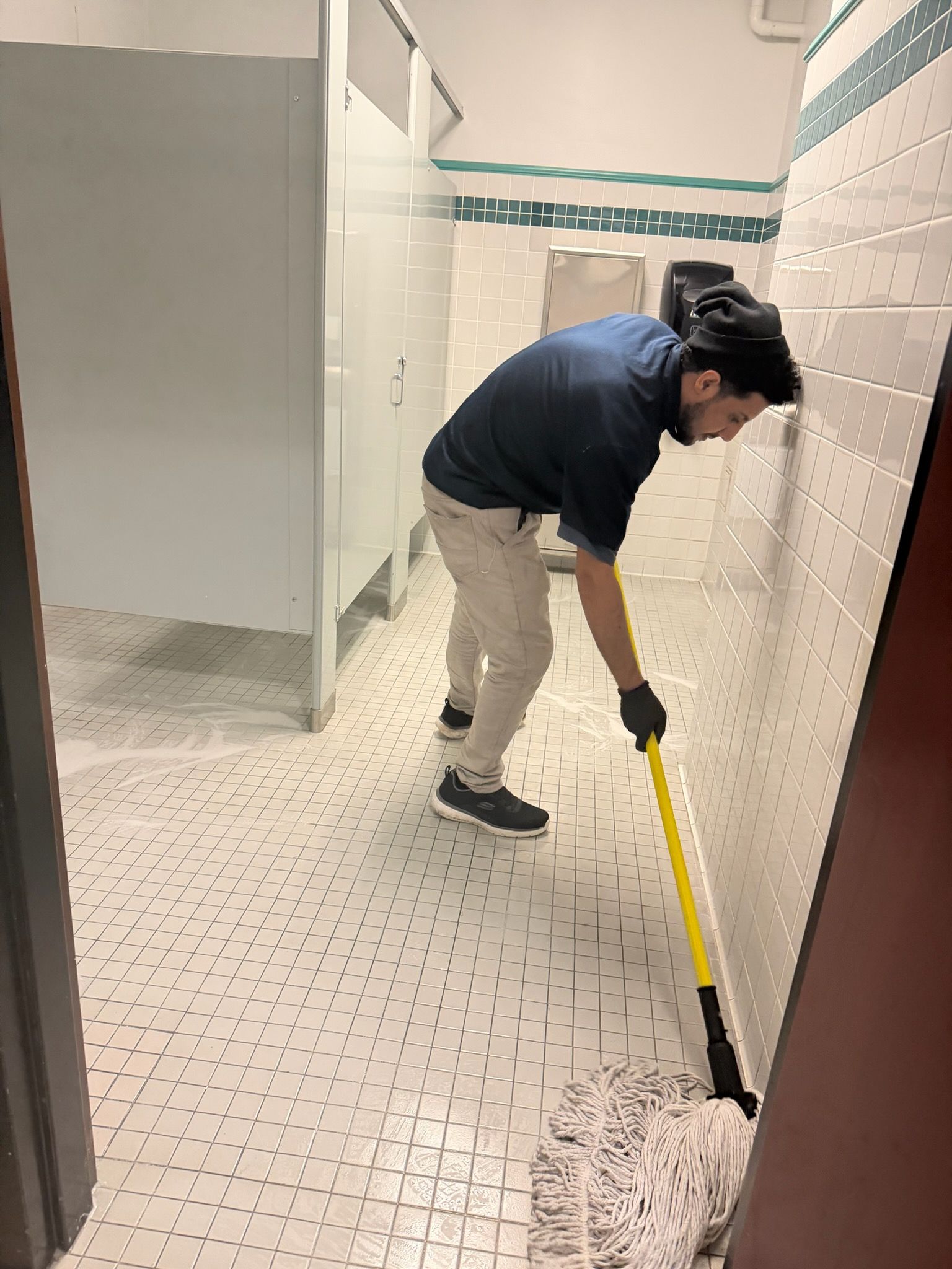 Person mopping a tiled restroom floor with a yellow mop handle
