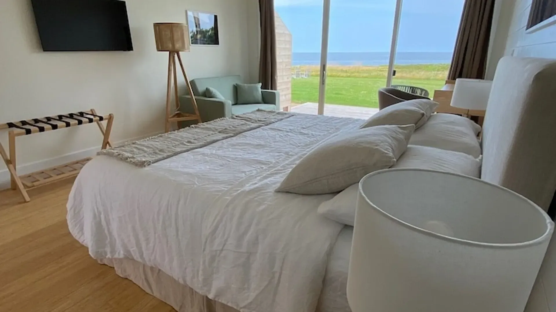 Lichte slaapkamer met een groot bed, staande lamp, stoel en uitzicht op de oceaan door glazen schuifdeuren.