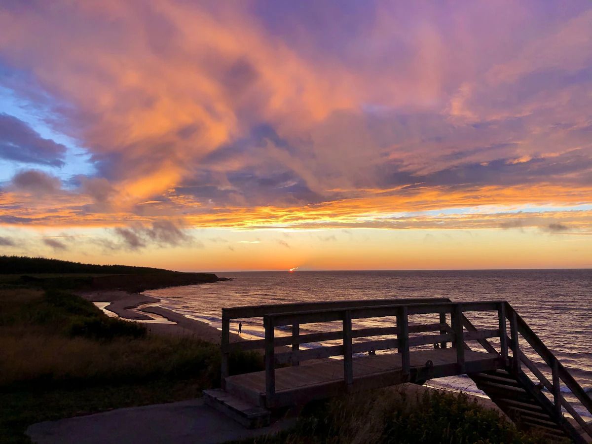 Zonsondergang boven een rotsachtige kustlijn met een houten uitkijkplatform en een kalme oceaan onder oranje en paarse wolken.