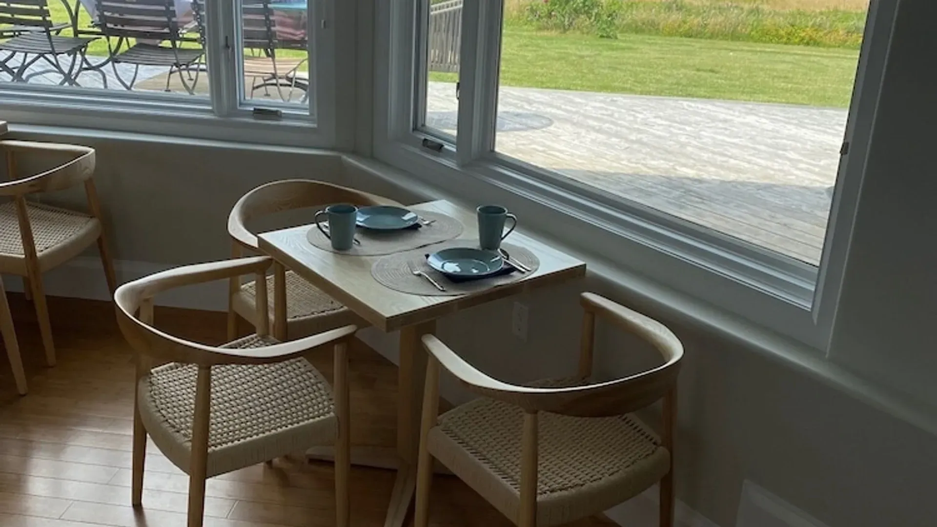 Twee houten stoelen aan een klein tafeltje bij een licht raam, met kopjes en schoteltjes, in een café.