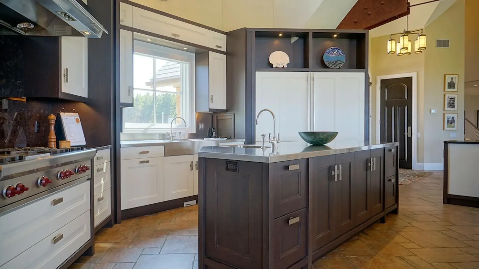 Ruime, moderne keuken met donker kookeiland, witte keukenkasten, groot raam en hanglampen.