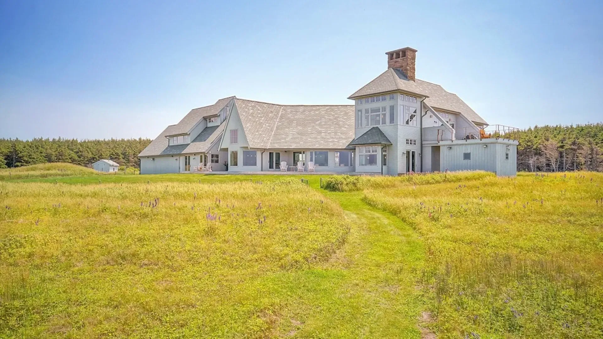 Een groot wit huis met een hoge schoorsteen staat in een grasveld onder een helderblauwe hemel.