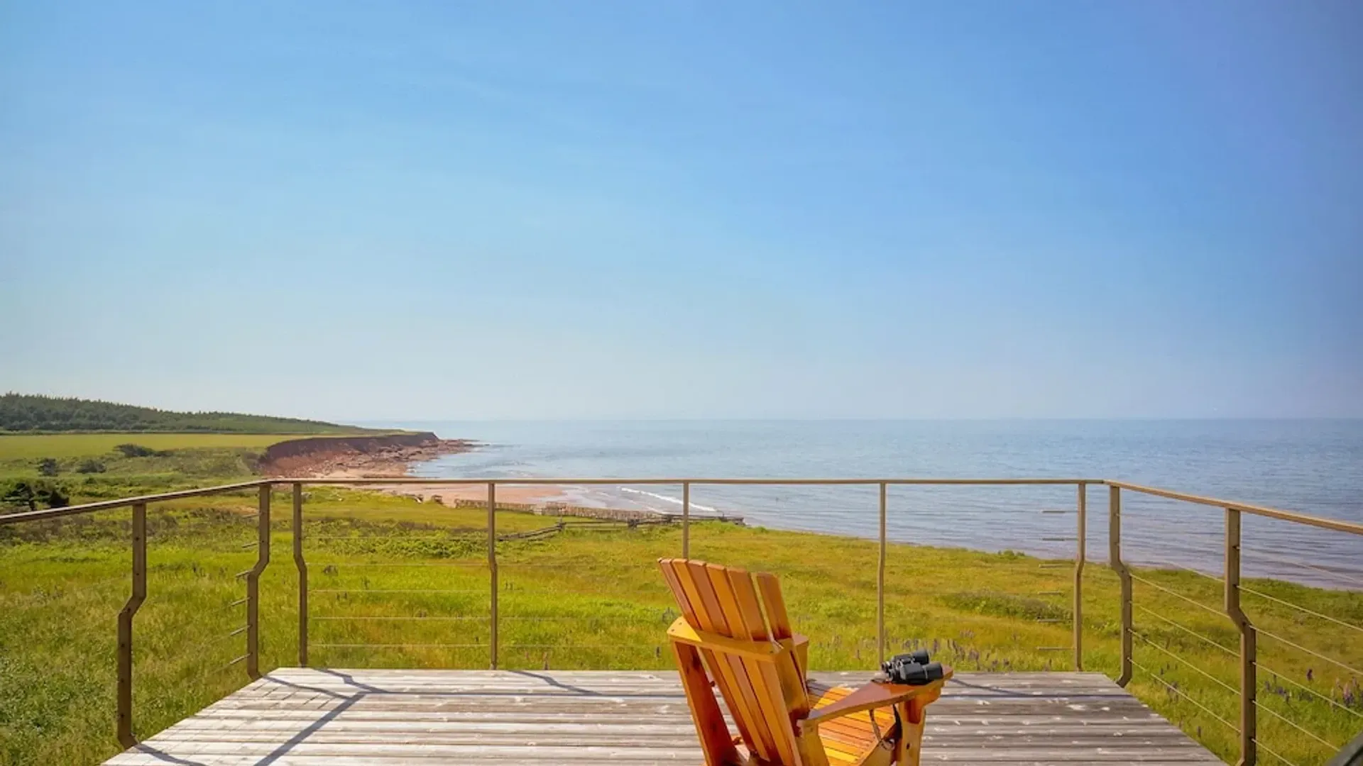 Zonnig terras aan zee met een houten stoel, met uitzicht op met gras begroeide kliffen en de oceaan.