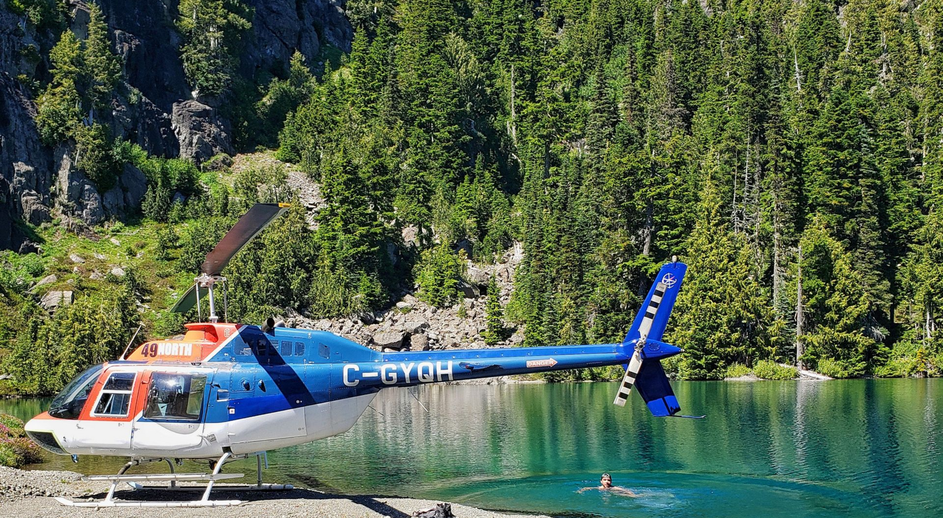 Een blauw-witte helikopter zweeft op een turquoise meer langs een beboste bergkust.