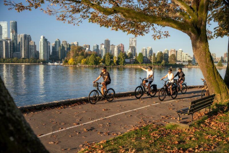 Fietsers rijden langs een met bomen omzoomd pad aan het water, met aan de overkant van het water de wolkenkrabbers van de stad op de achtergrond bij zonsondergang.