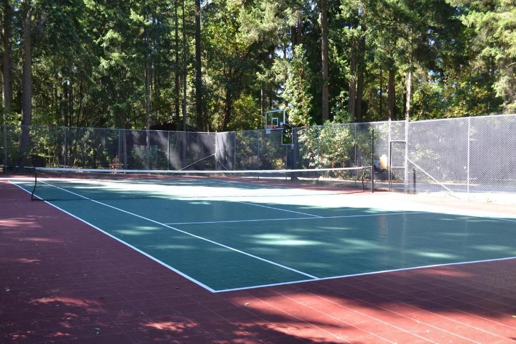 Een tennisbaan in de openlucht met een groen speeloppervlak en een rode rand, omgeven door bomen en een hek.
