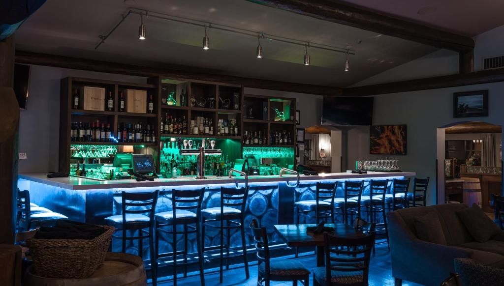 Een schemerig verlichte bar met een blauwgroen verlichte toonbank, schappen vol flessen en lege tafels en stoelen.