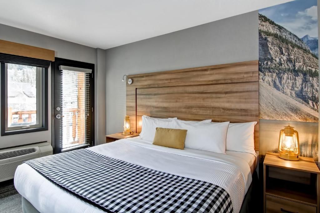 Moderne slaapkamer met een groot bed, houten hoofdbord, nachtlampjes en een kunstwerk met bergmotief bij het raam.