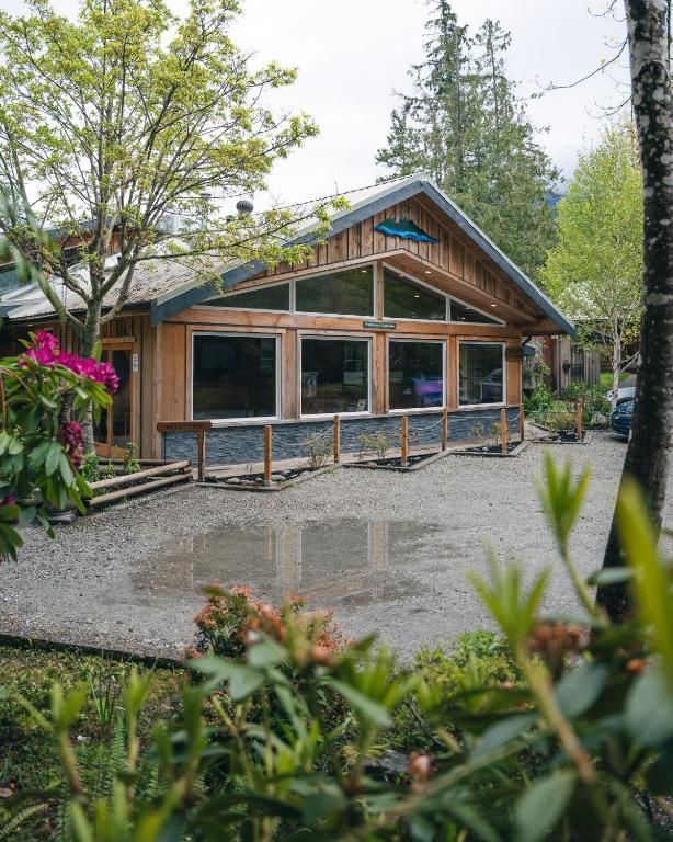 Rustieke blokhut met grote ramen en een stenen fundering, omgeven door bomen en een grindhof na regen.