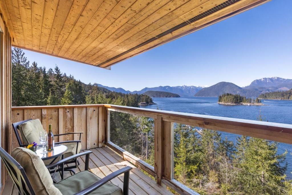 Overdekt terras met uitzicht op een blauw meer en beboste bergen, met twee stoelen en een houten balustrade.