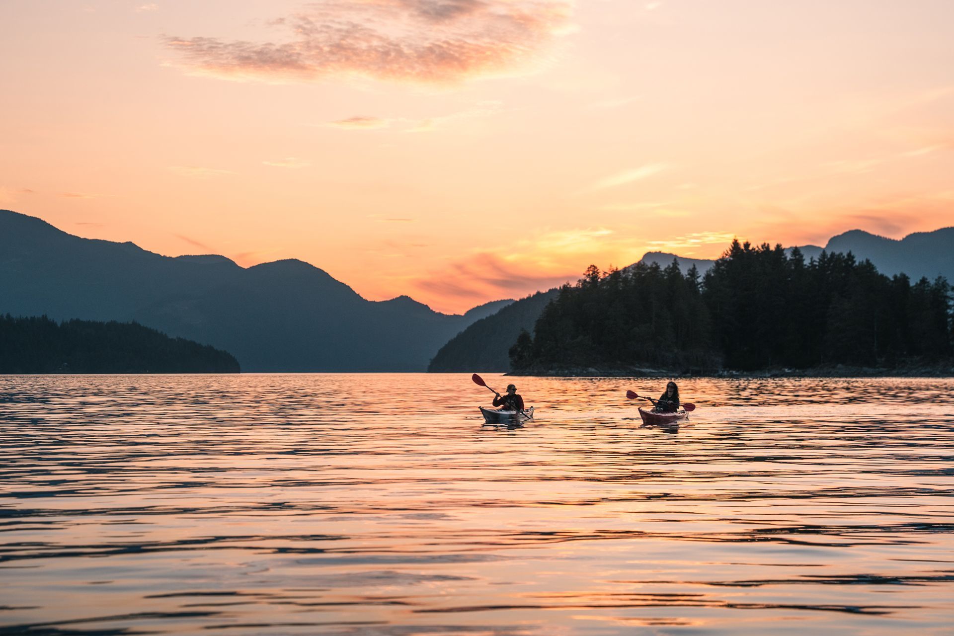 Kajakkers op een kalm meer bij zonsondergang met bergen en een beboste oever op de achtergrond.