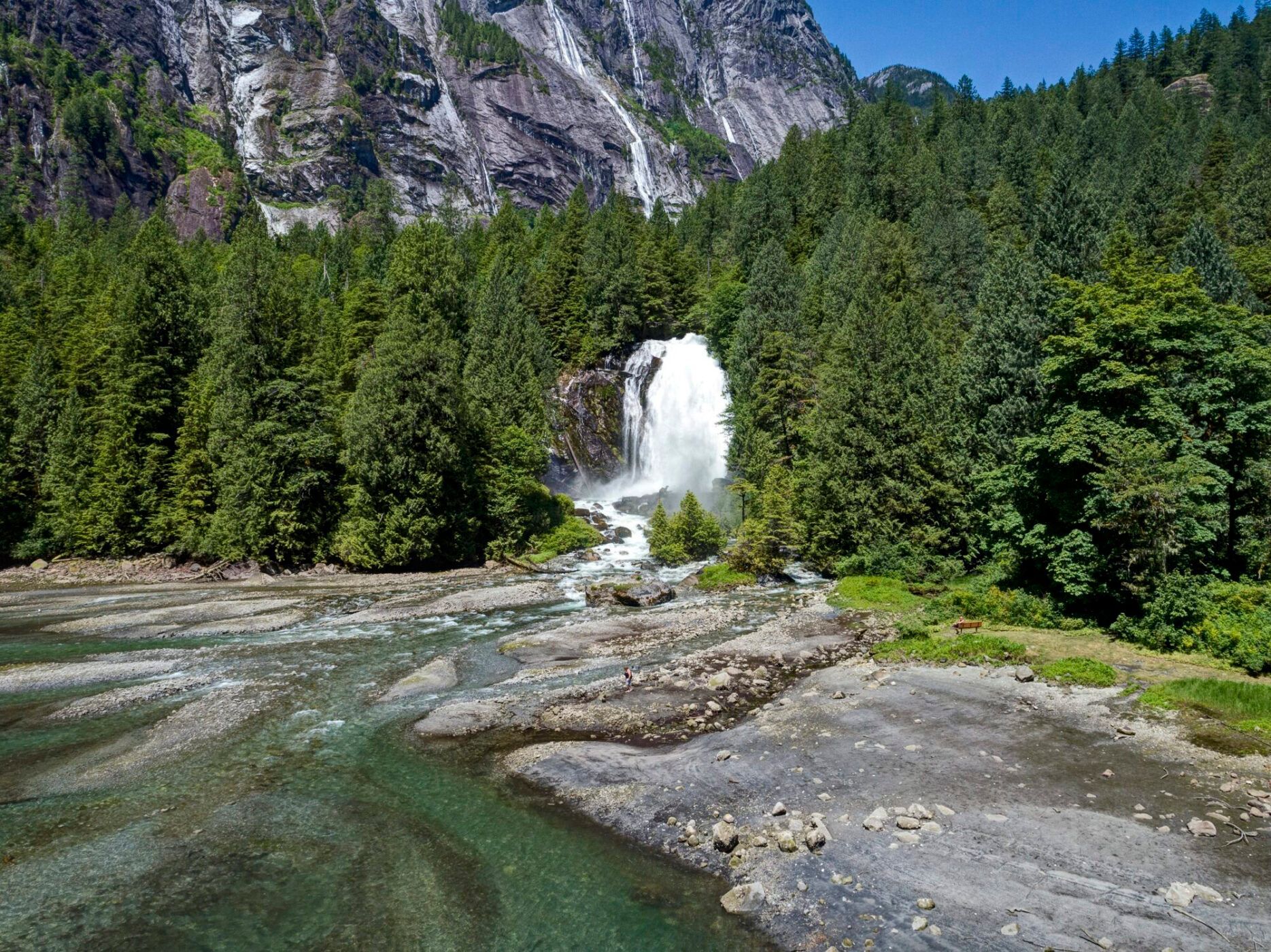 Waterval in een bebost bergdal naast een rotsachtige rivieroever onder een stralende zon.
