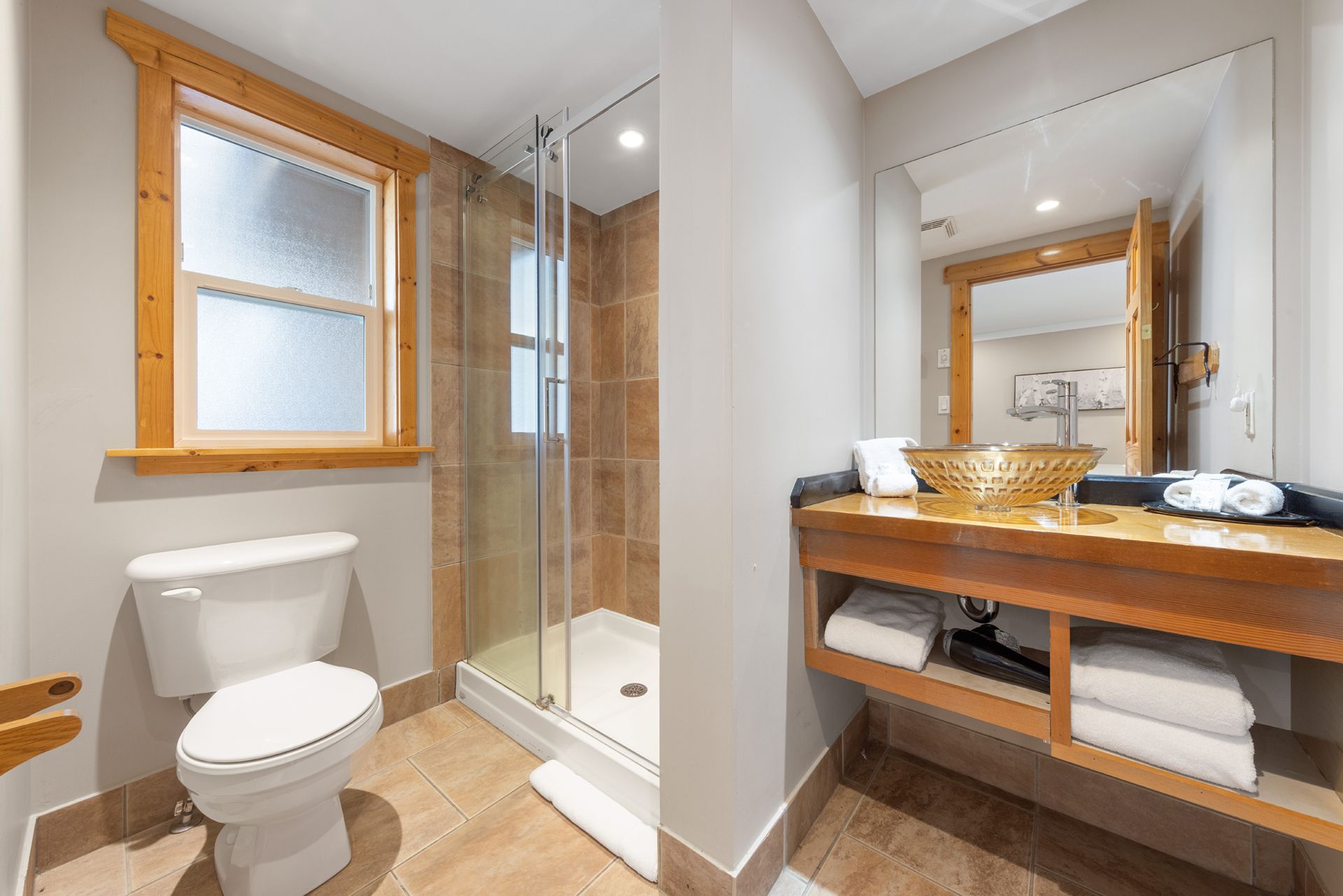 Moderne badkamer met glazen douchecabine, toilet, houten wastafelmeubel en spiegel bij het raam.