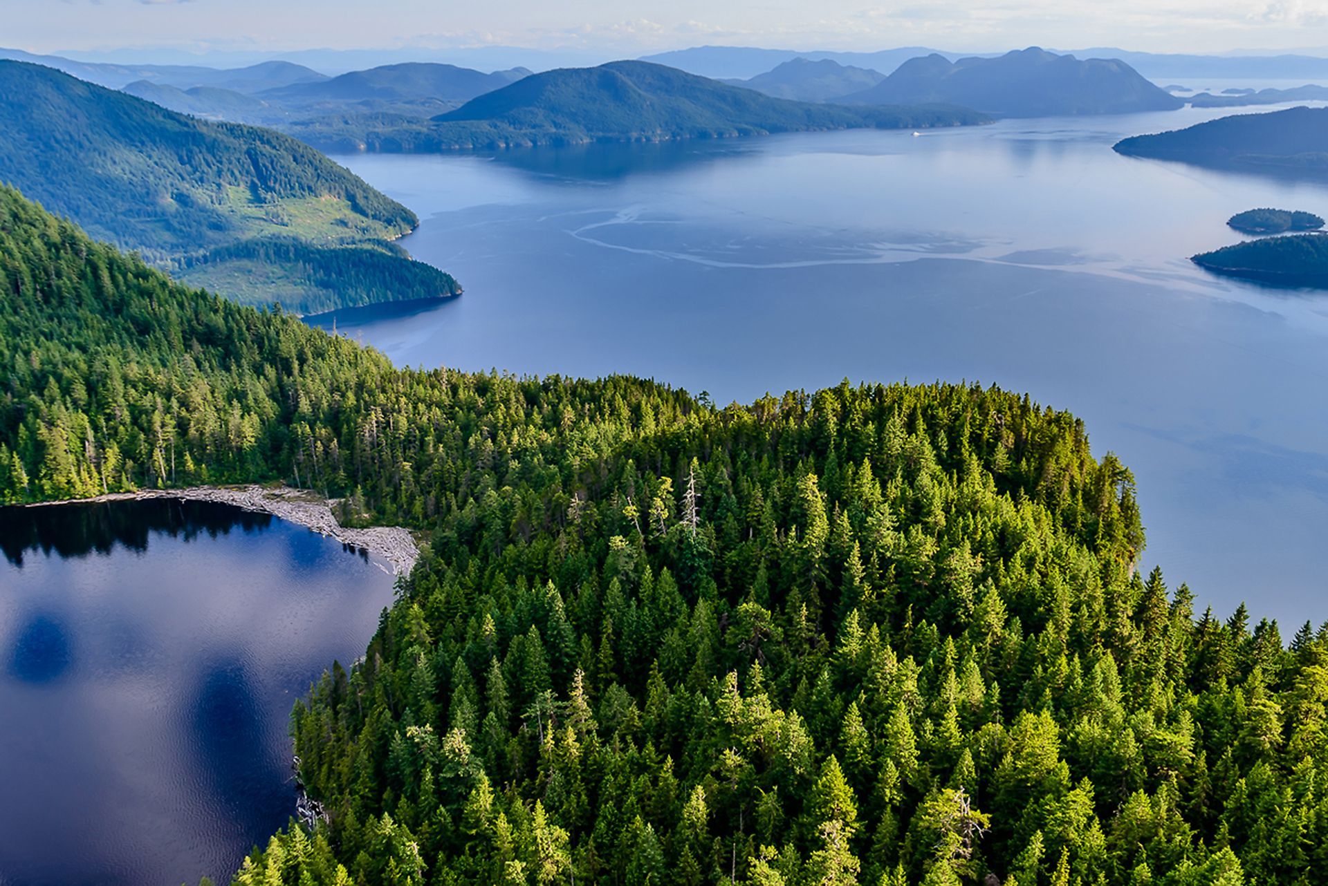 Luchtfoto van een bebost schiereiland en een blauw meer met in de verte bergen onder een heldere hemel.