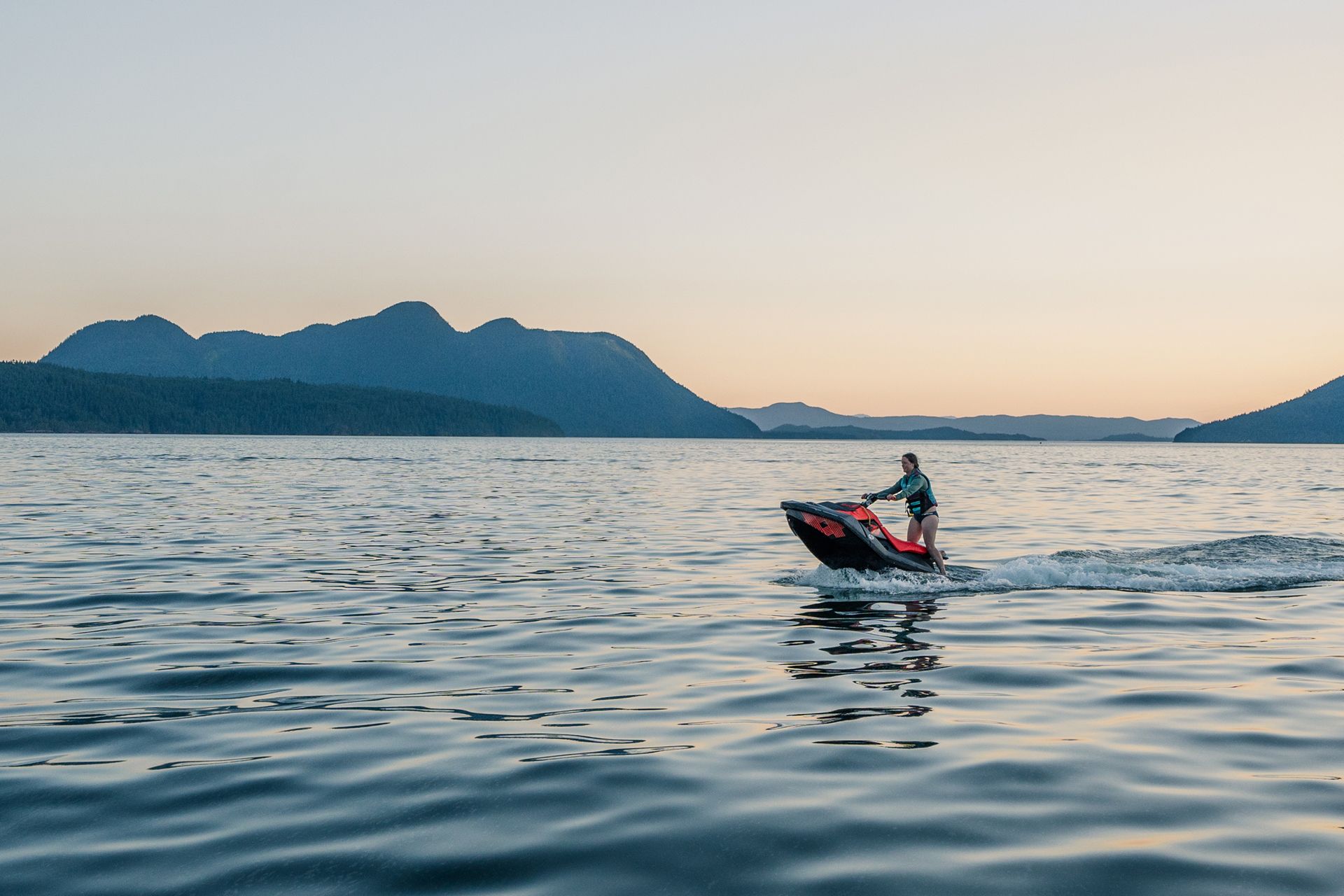 Een persoon vaart op een jetski op kalm water bij zonsondergang, met bergen op de achtergrond.