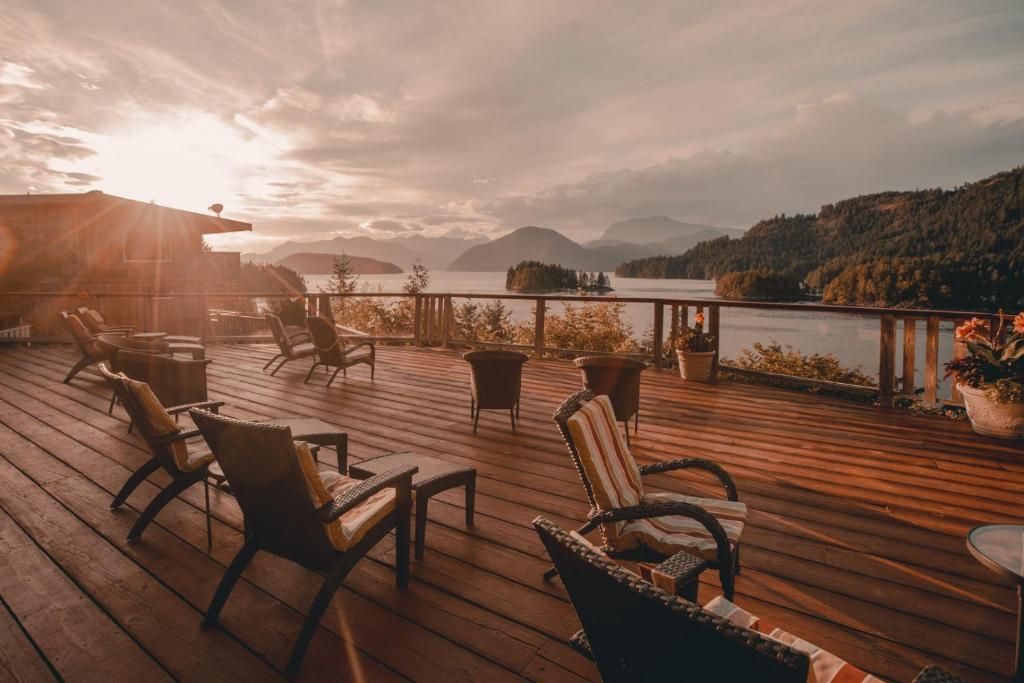 Houten terras met ligstoelen, met uitzicht op een rustig meer bij zonsondergang, met bergen en een warme oranje lucht.