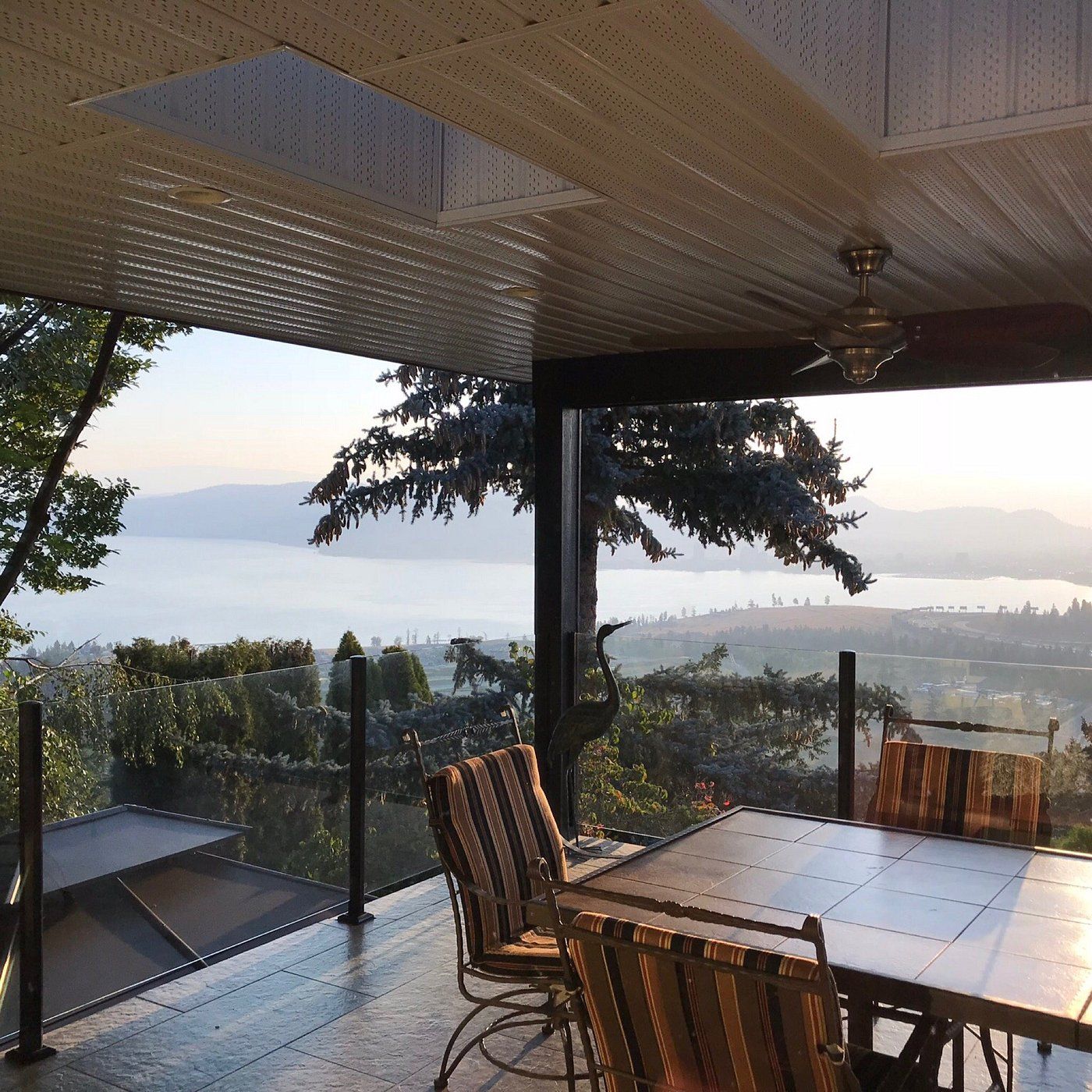 Overdekt terras met glazen balustrade, eettafel en stoelen, met uitzicht op een mistig meer en de bergen bij zonsondergang.