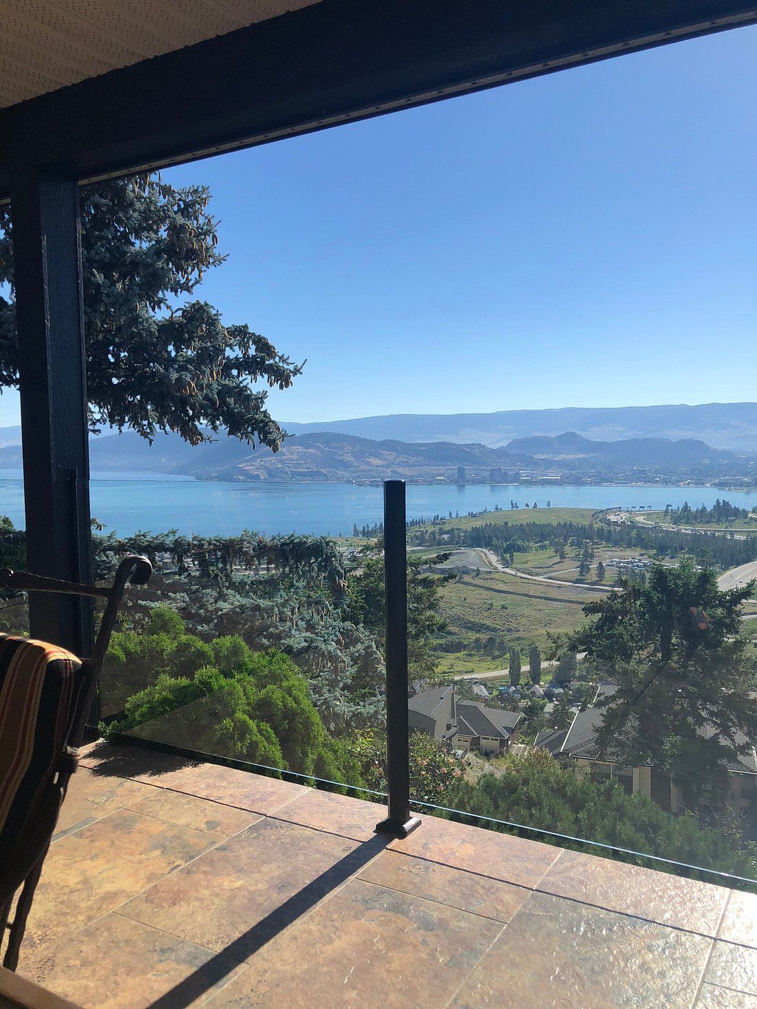 Zonnig terras met uitzicht op een baai en heuvels, omlijst door een glazen balustrade en boomtakken.