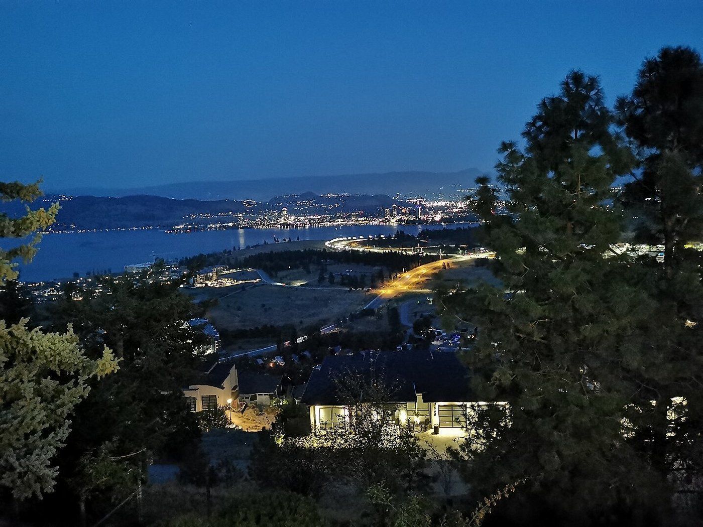 Een kustlandschap in de schemering met gloeiende lichtjes, donkere heuvels en bomen die het uitzicht op de baai omlijsten.