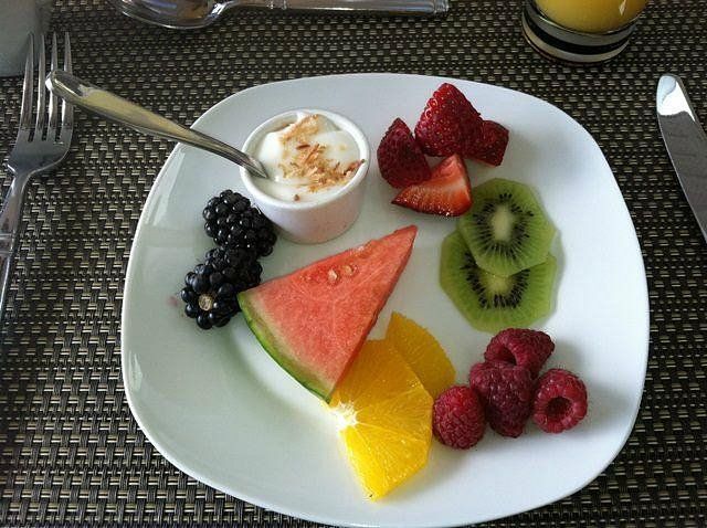 Een fruitschaal met yoghurtdip, bessen, kiwi, watermeloen en sinaasappelschijfjes op tafel.