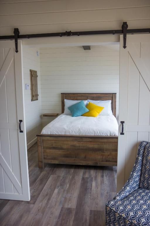 Rustieke slaapkamer met een houten bed, wit beddengoed en gele sierkussens achter schuifdeuren in boerderijstijl.