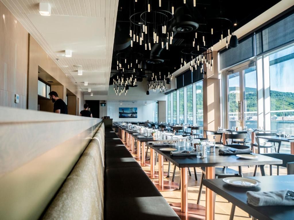 Moderne restaurantzaal met lange tafels, zwarte plafondlampen en grote ramen met uitzicht op het water.