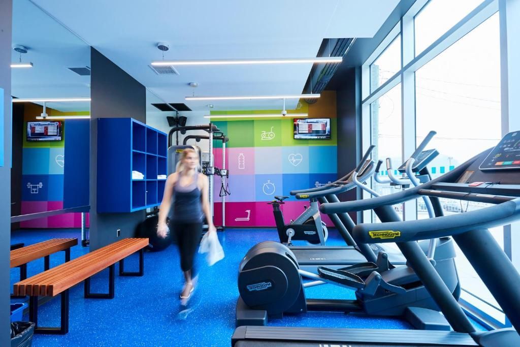 Moderne sportschool met loopbanden, blauwe vloer, kleurrijke kluisjes en een persoon die bij de bankjes loopt.
