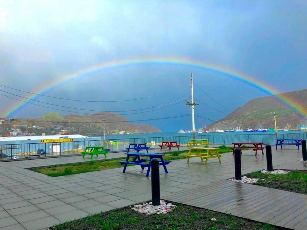 Een regenboog boven een promenade langs het water met kleurrijke bankjes, een hoog beeldhouwwerk en mistige heuvels op de achtergrond.