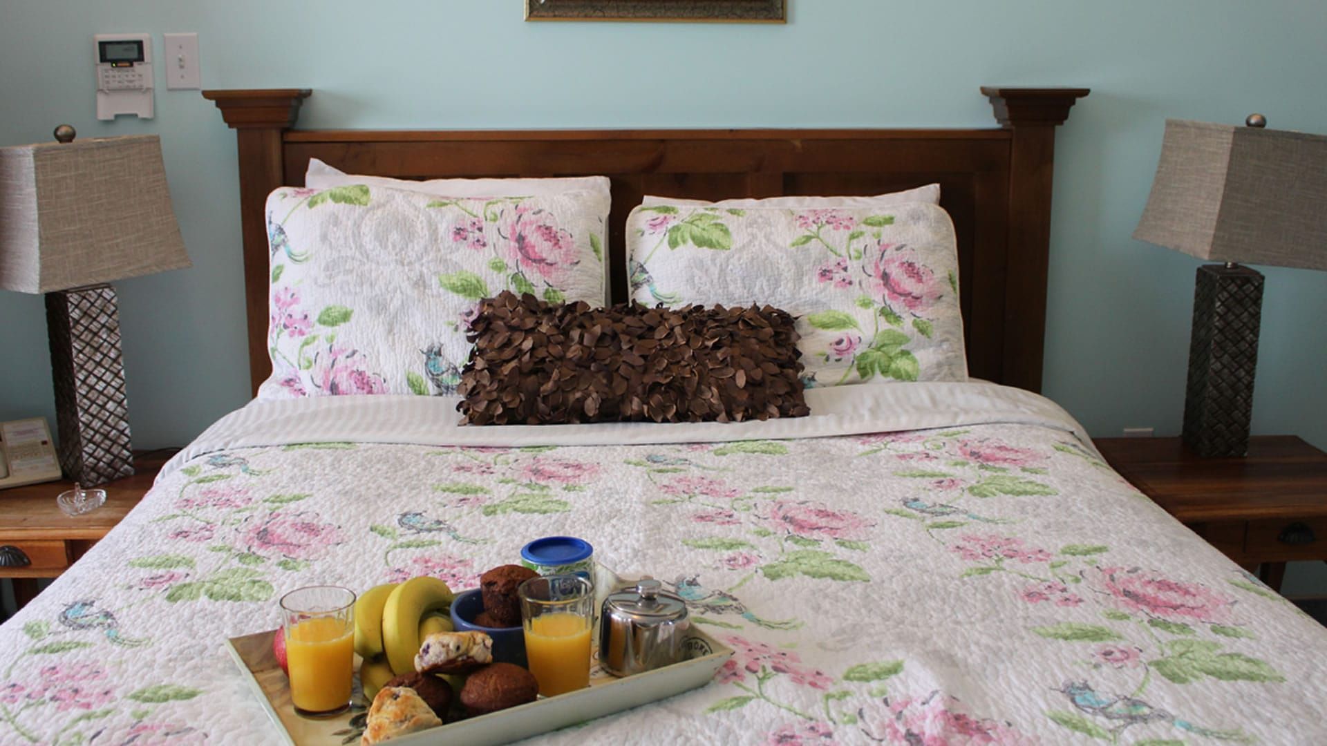 Netjes opgemaakt bed met bloemenbeddengoed, twee nachtlampjes en een ontbijtplateau met fruit aan het voeteneinde van het bed.