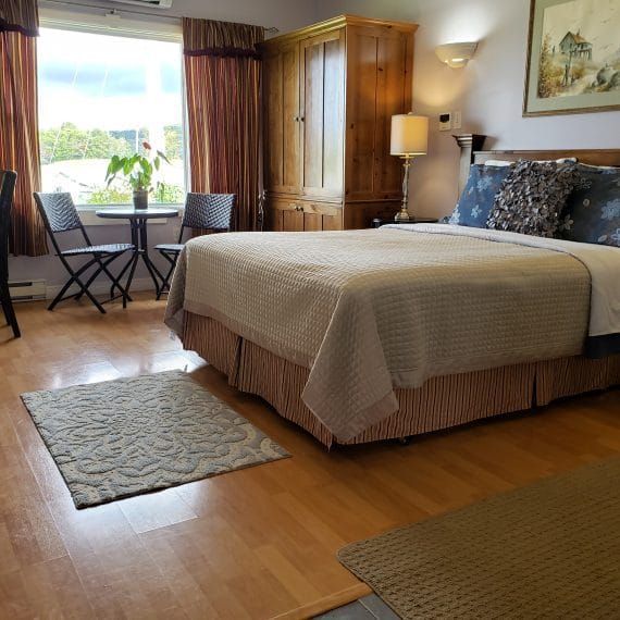 Lichte slaapkamer met een groot bed, houten kledingkast, tafeltje bij het raam en vloerkleden op een houten vloer.